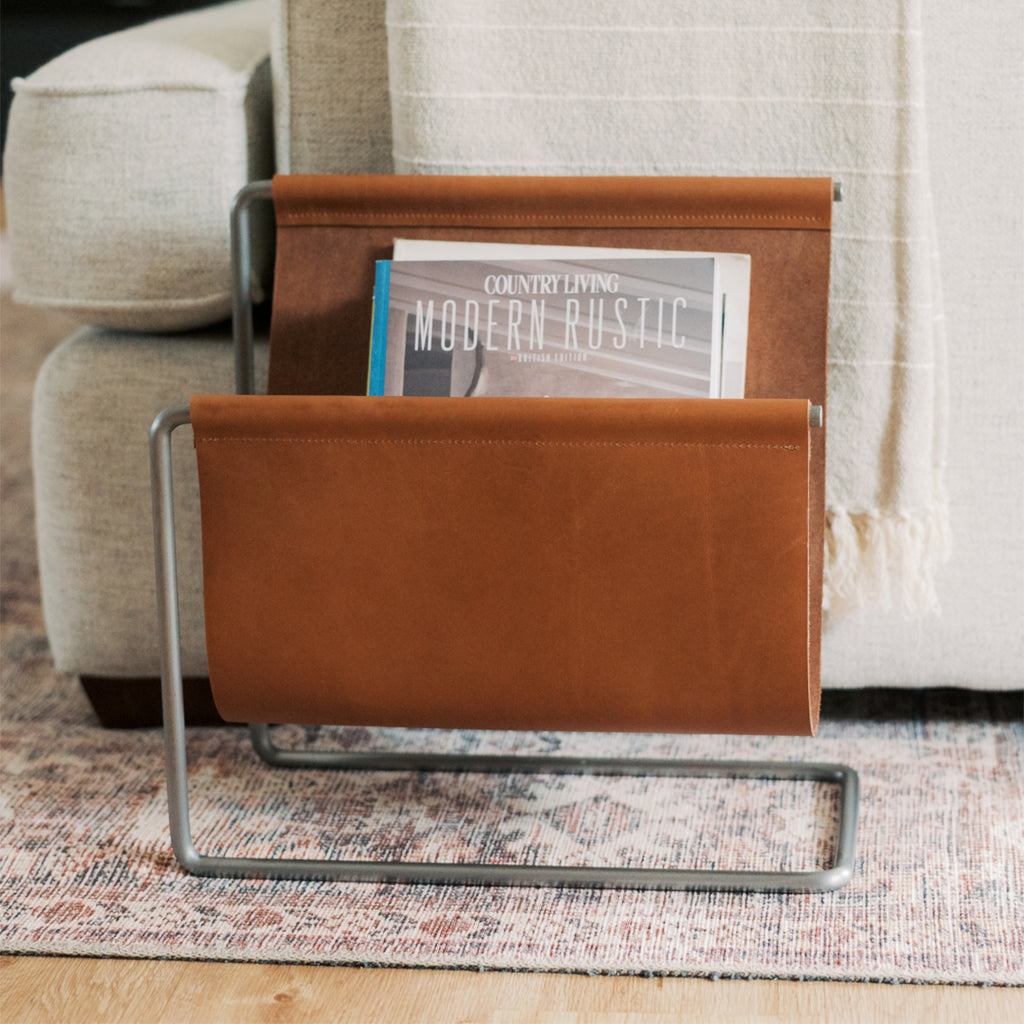 Tan full-grain leather magazine rack with handmade steel frame, holding magazines beside a sofa in a cozy living room setting