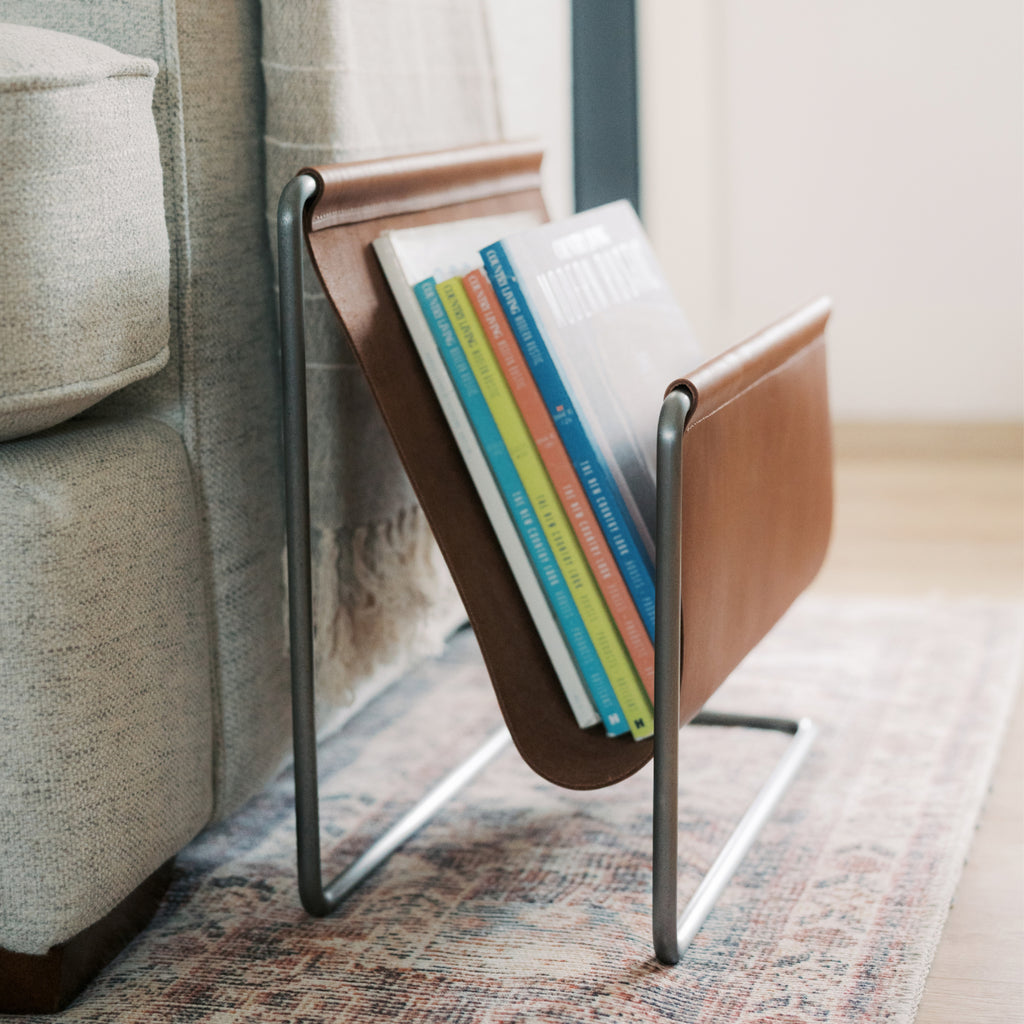 Tan full-grain leather magazine rack with handmade steel frame, holding magazines beside a sofa in a cozy living room setting