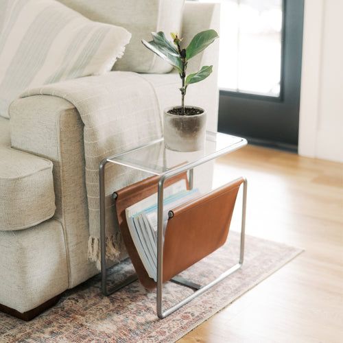 Modern steel and leather magazine rack side table with tempered glass top and full-grain leather sling, styled beside a sofa in a bright living room