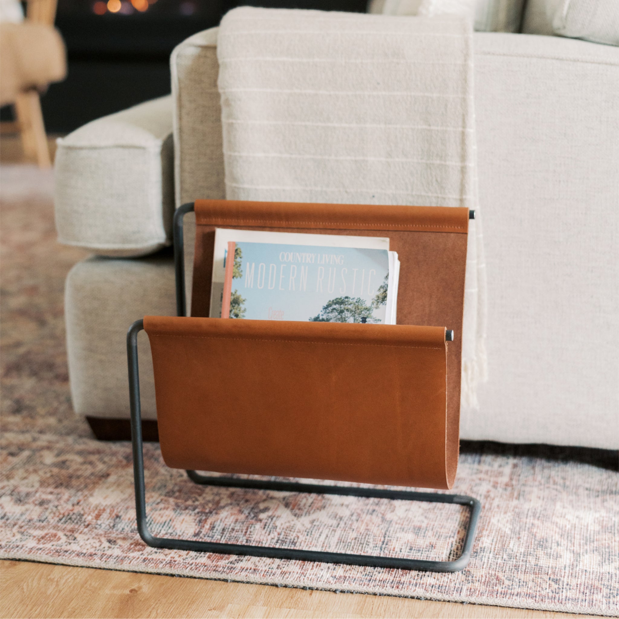 Tan full-grain leather magazine rack with handmade dark steel frame, holding magazines beside a sofa in a cozy living room setting
