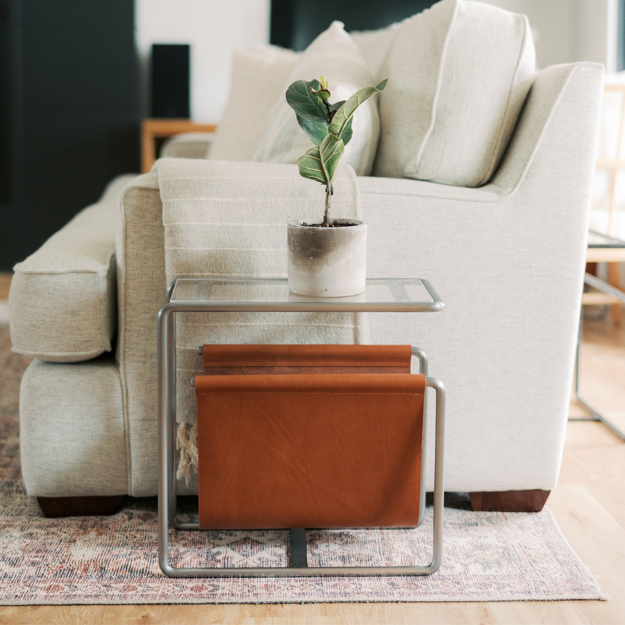 Raw steel and leather magazine rack side table with tempered glass top, caramel leather sling, and handmade steel frame in a modern living room setting